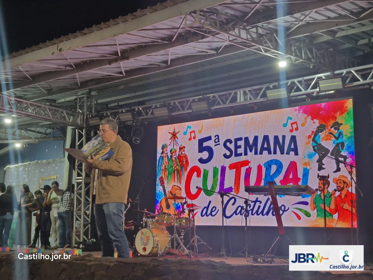Apresentador ao microfone diante do painel da quinta Semana Cultural de Castilho