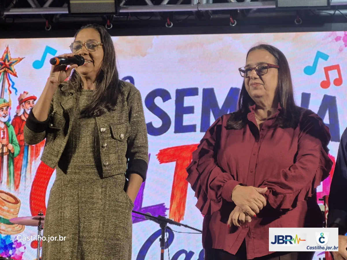 Mulher discursa ao microfone ao lado de outra participante no palco
